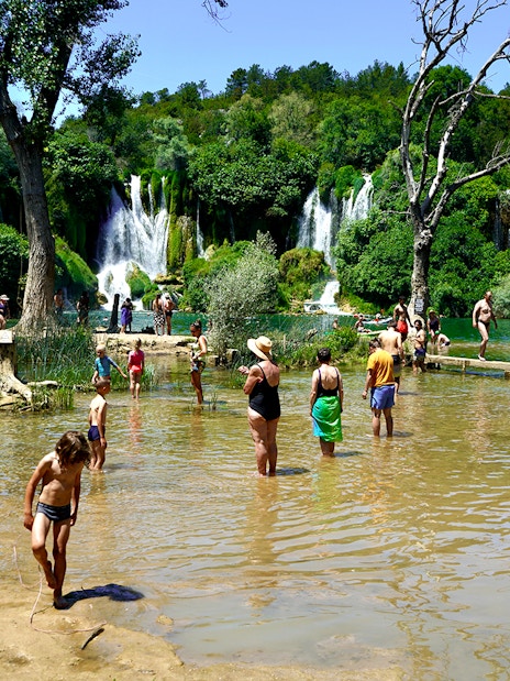 Tourists wading in water at Kravica Falls, Bosnia, with lush greenery and waterfalls in the background.