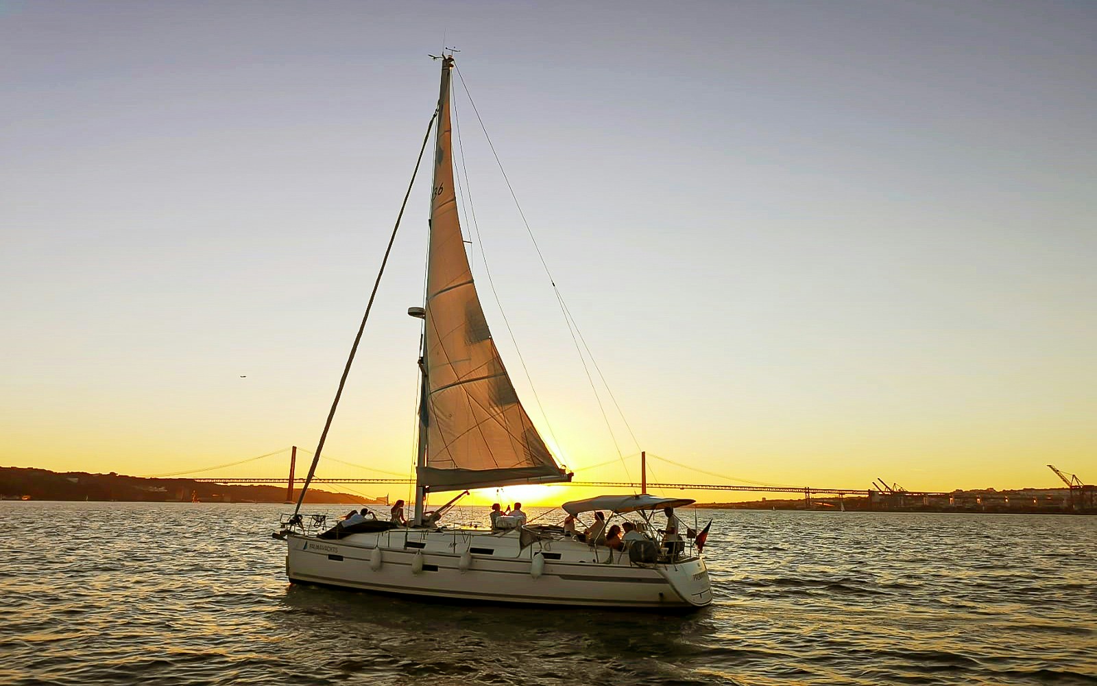 Sailboat on Tagus River at sunset with Lisbon's 25 de Abril Bridge in the background.