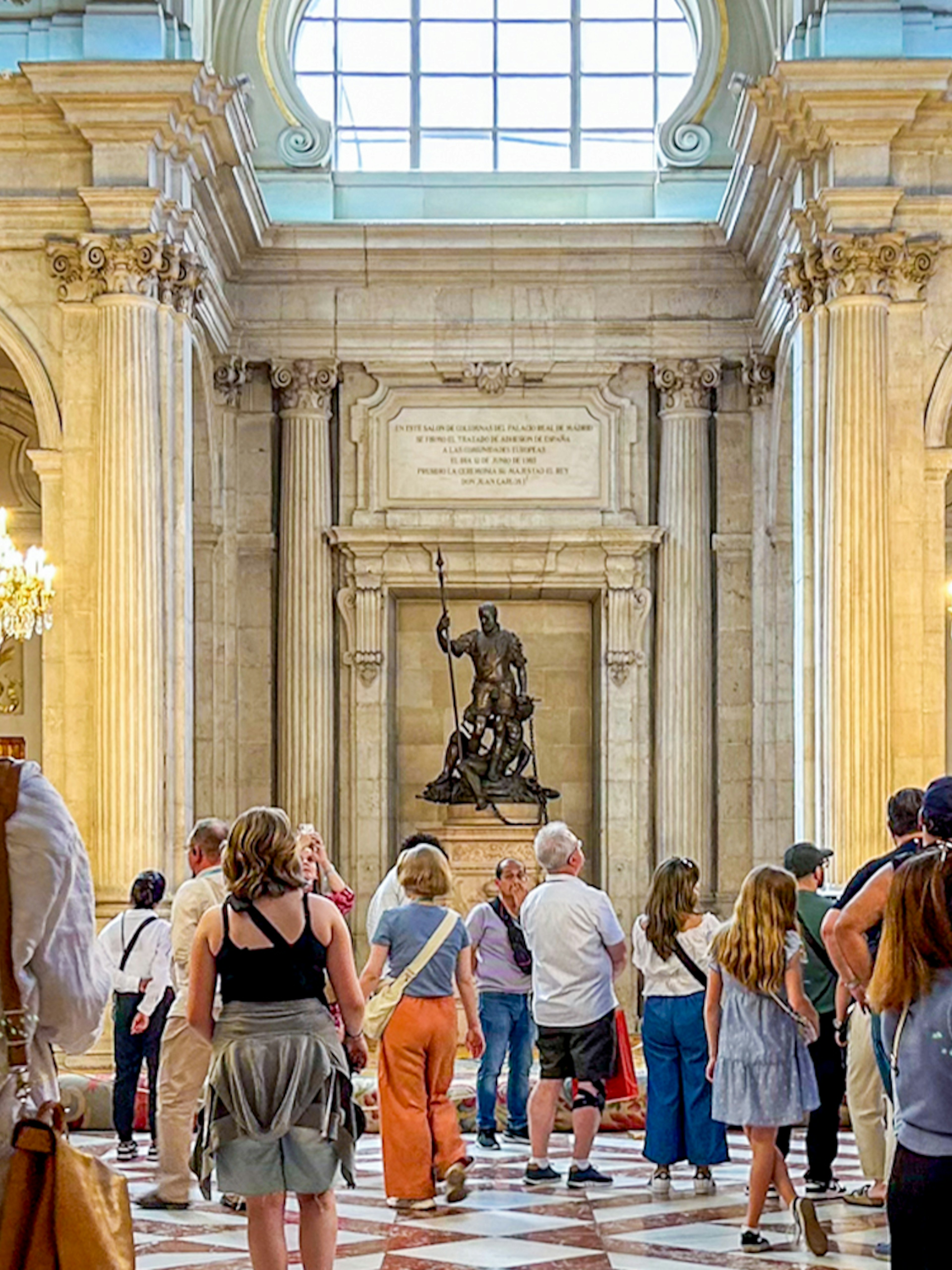 Visitors exploring the ornate hall of the Royal Palace of Madrid with chandeliers and statues.