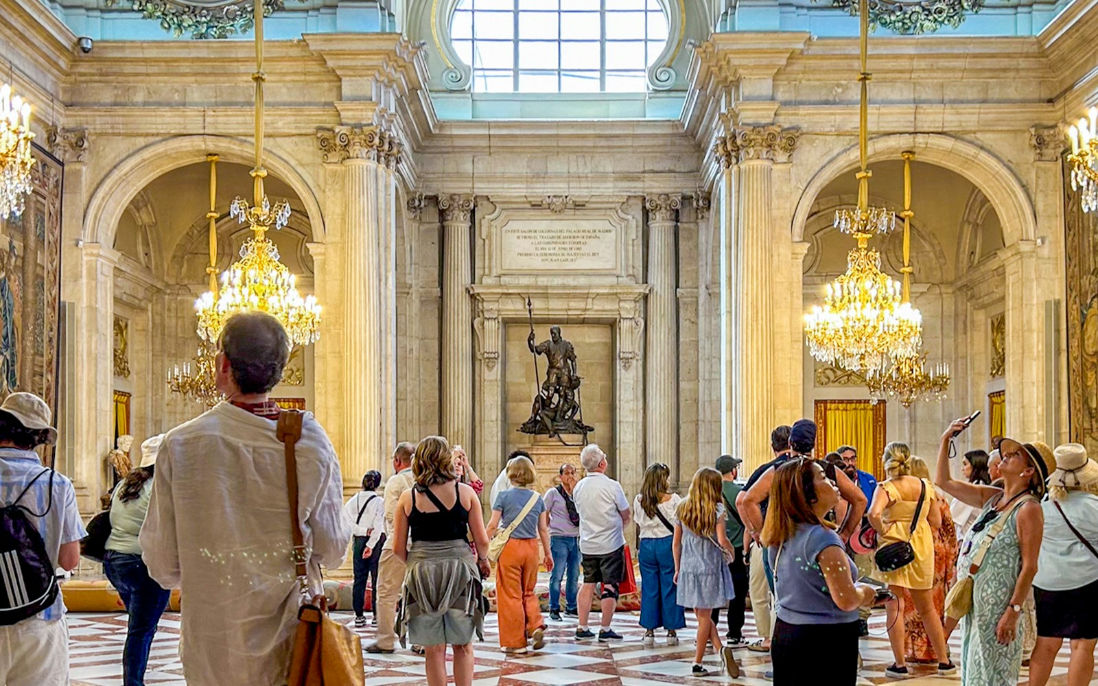 Visitors exploring the ornate hall of the Royal Palace of Madrid with chandeliers and statues.