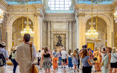 Visitors exploring the ornate hall of the Royal Palace of Madrid with chandeliers and statues.