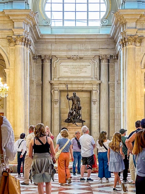 Visitors exploring the ornate hall of the Royal Palace of Madrid with chandeliers and statues.