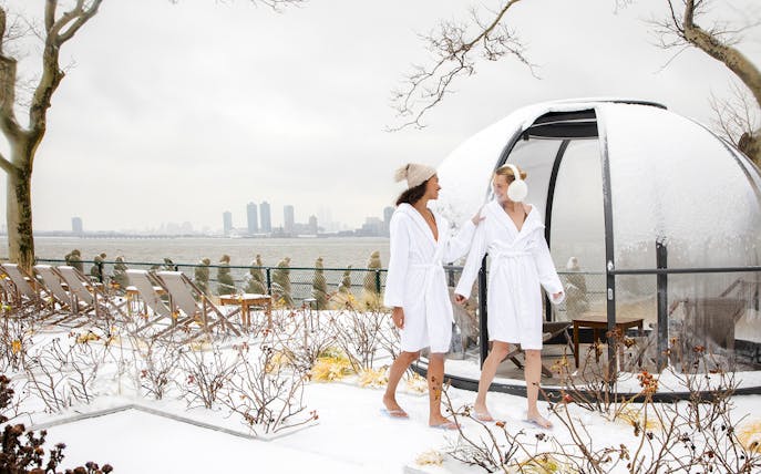 Two people in robes walking on a snowy terrace at QC New York spa with city skyline in the background.