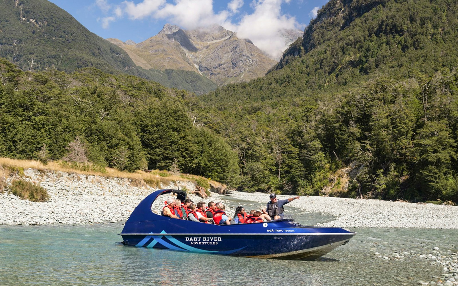 Jet boat on Dart River with tourists, surrounded by lush forest and mountains, New Zealand.