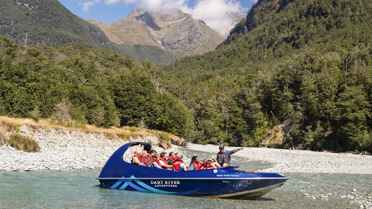 Jet boat on Dart River with tourists, surrounded by lush forest and mountains, New Zealand.