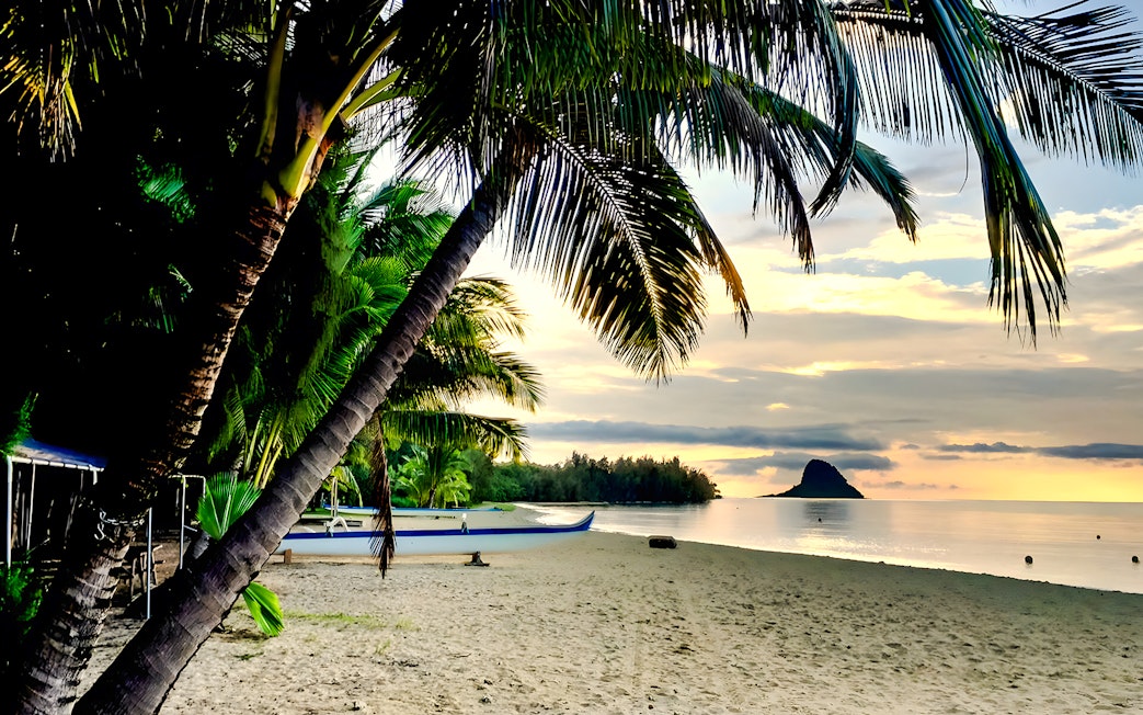 Secret Island Beach at sunset with palm trees and a canoe, Kualoa Ranch, Hawaii.