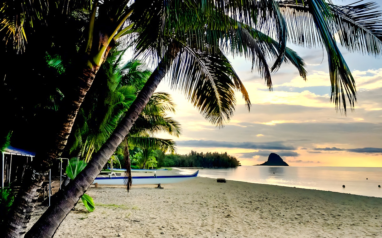 Secret Island Beach at sunset with palm trees and a canoe, Kualoa Ranch, Hawaii.