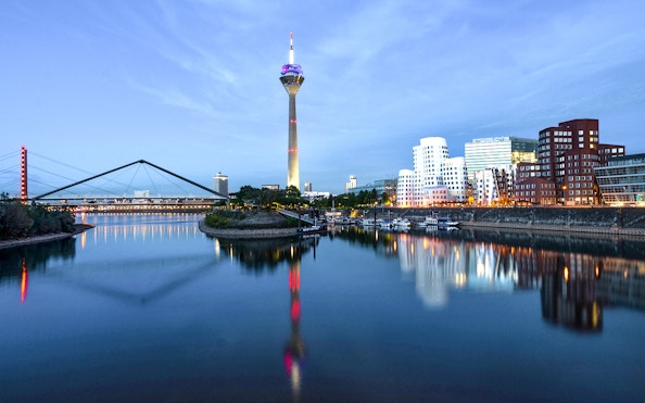 Evening view of Düsseldorf skyline with Rheinturm and bridge reflecting in the Rhine River.