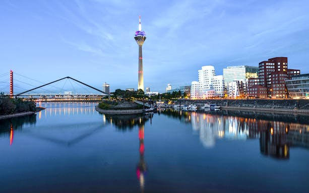 Evening view of Düsseldorf skyline with Rheinturm and bridge reflecting in the Rhine River.