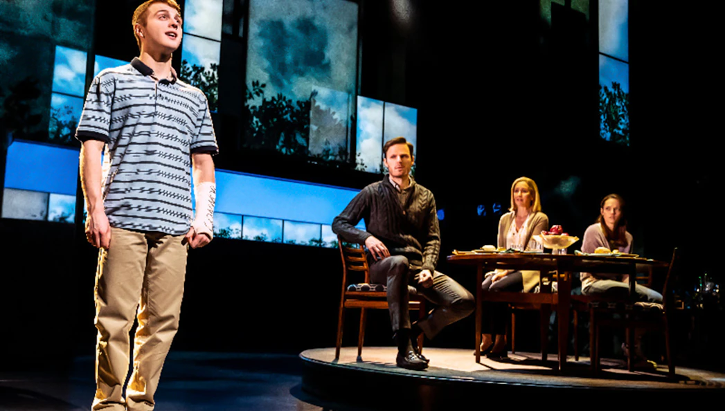 Stage scene from "Dear Evan Hansen" with a young man standing and three people seated at a table.