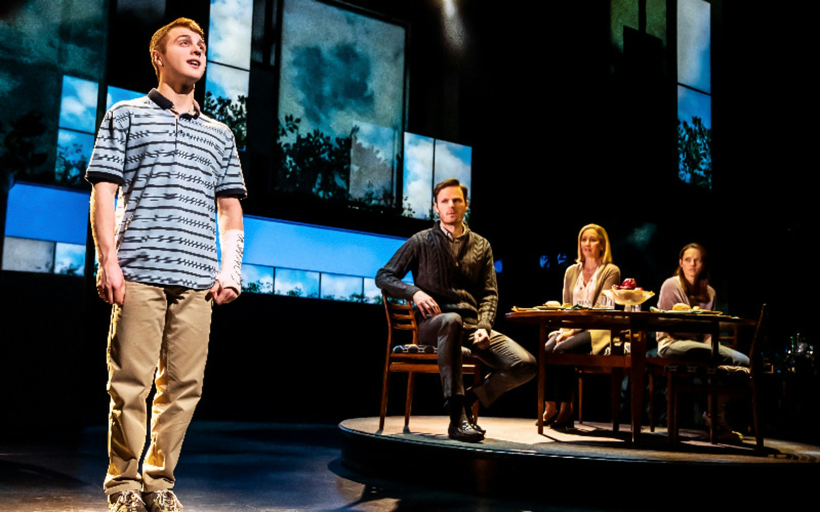 Stage scene from "Dear Evan Hansen" with a young man standing and three people seated at a table.