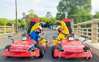 Go-kart riders in costumes pose on a street in Osaka Namba with Osaka Castle in the background.