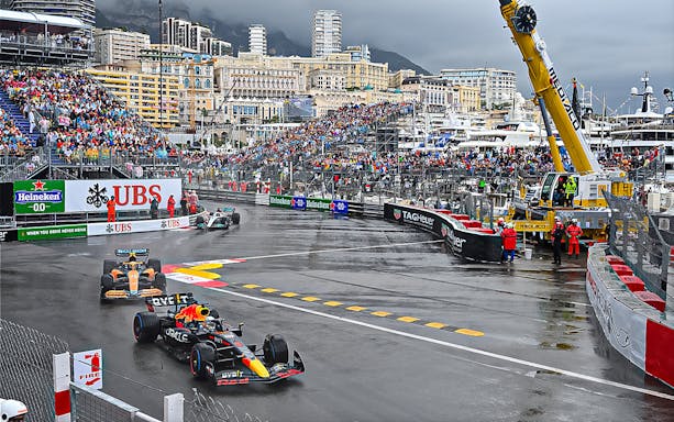 Formula 1 cars racing on a wet track at Monaco Grand Prix with crowded stands.