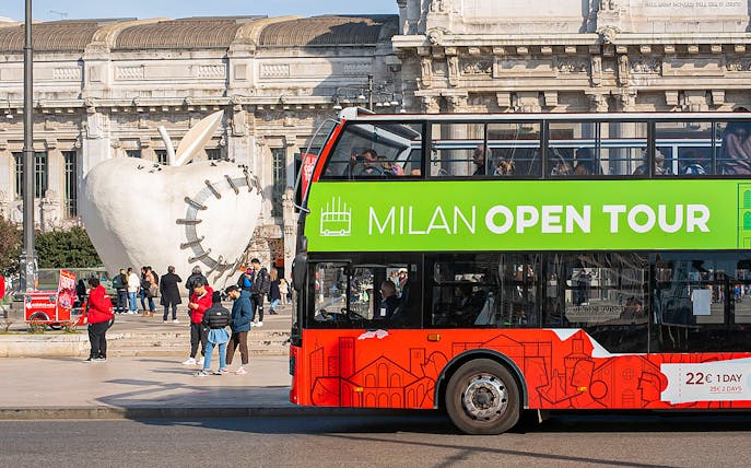 Hop-on hop-off bus near Milan's Central Station with apple sculpture in background.