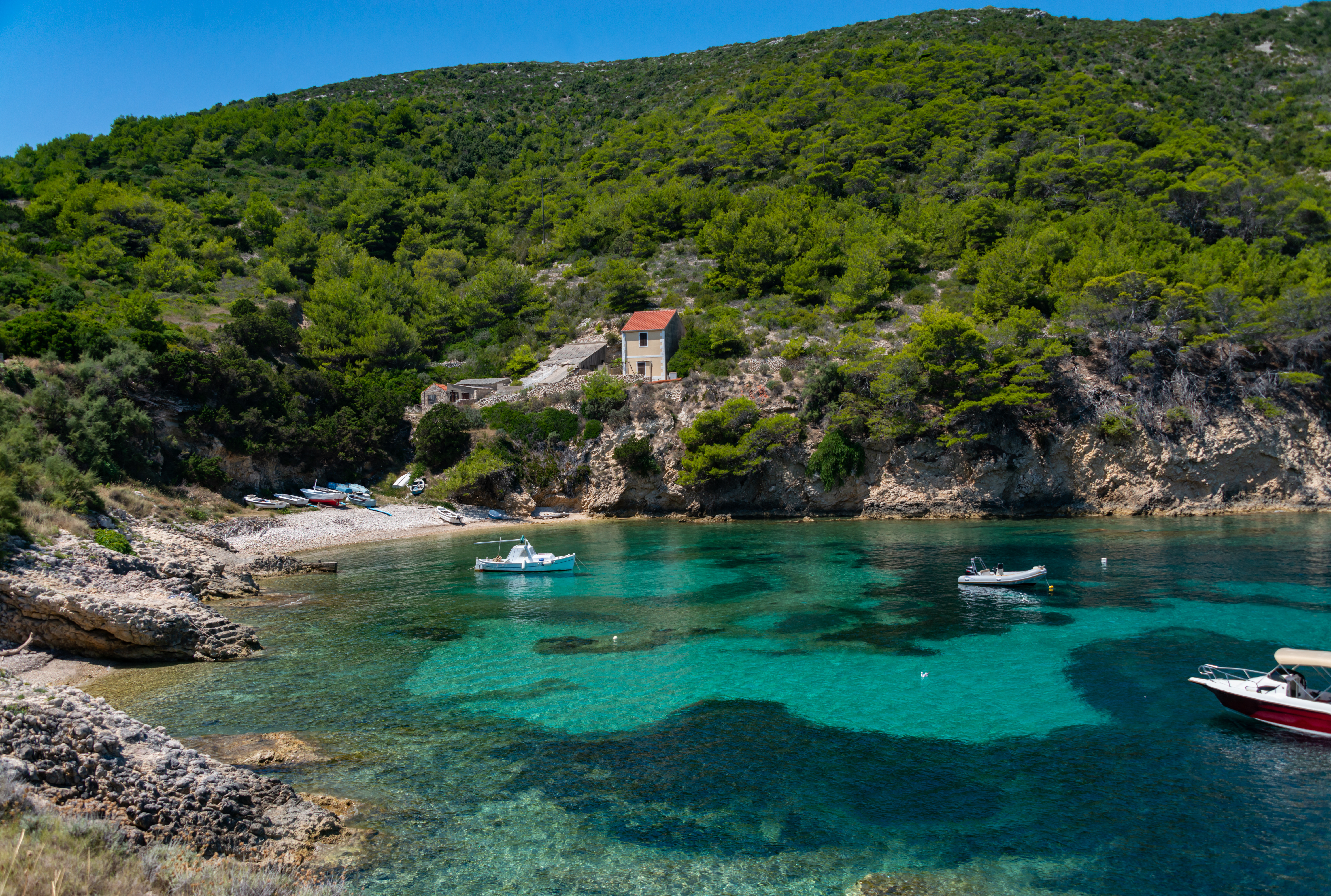 Boats anchored in a clear turquoise bay with a hillside and house on Biševo Island, Adriatic Sea.