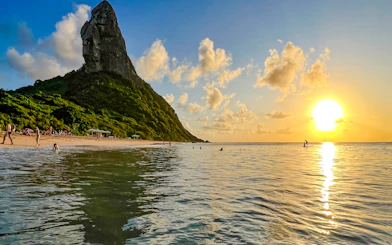 Sunset over beach with Morro do Pico, people swimming and paddleboarding, Fernando de Noronha.