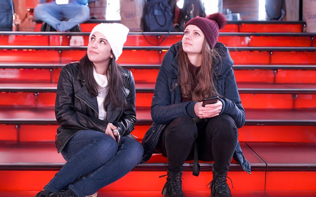 Two girls sitting on the red steps at Times Square at night.