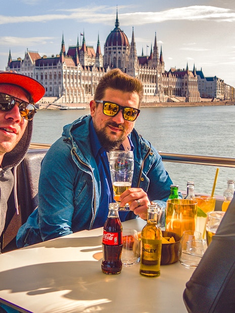 Group enjoying drinks on a Budapest party boat cruise with the Hungarian Parliament in the background.