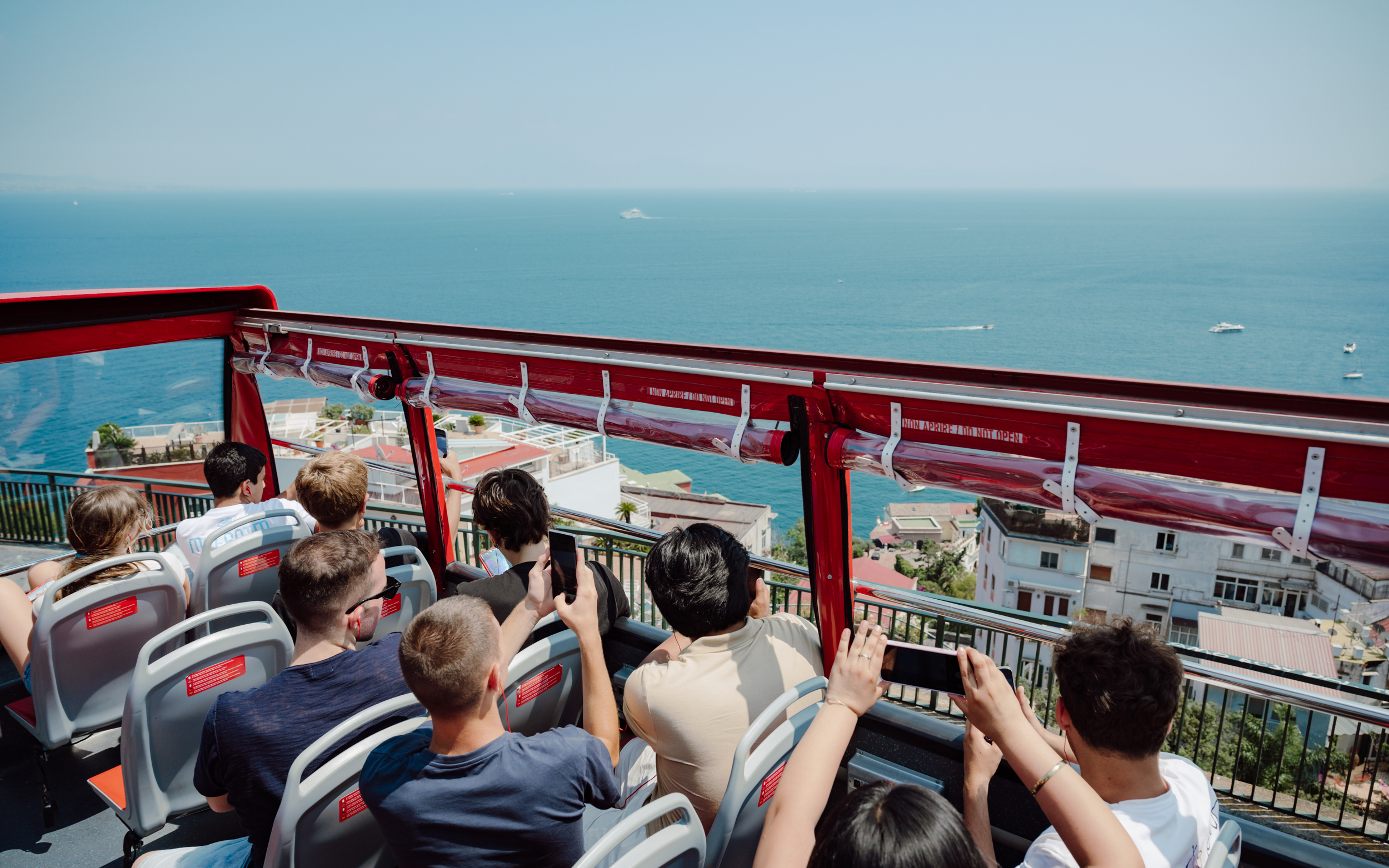 Tourists on open-top bus enjoying Naples coastline view.