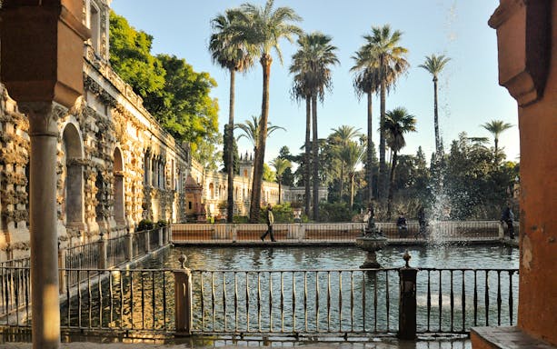 Alcazar Seville gardens with palm trees and fountain by historic stone walls.