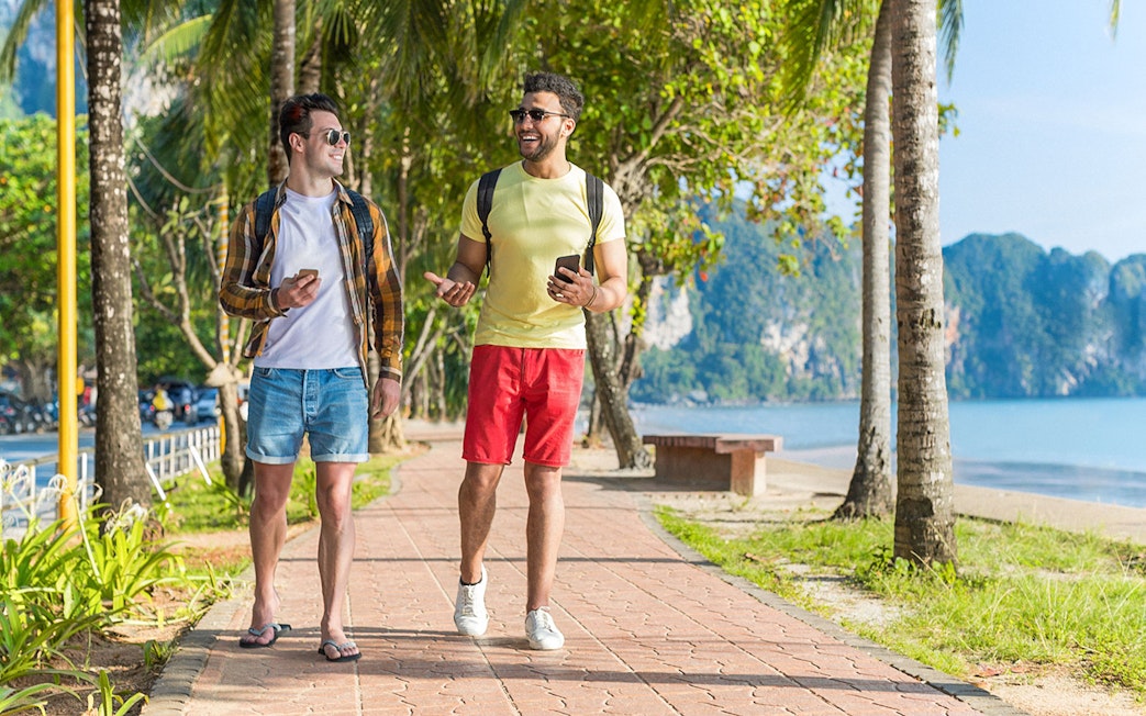 Two people walking on a palm-lined path by the beach, using phones in Kuala Lumpur, Malaysia.