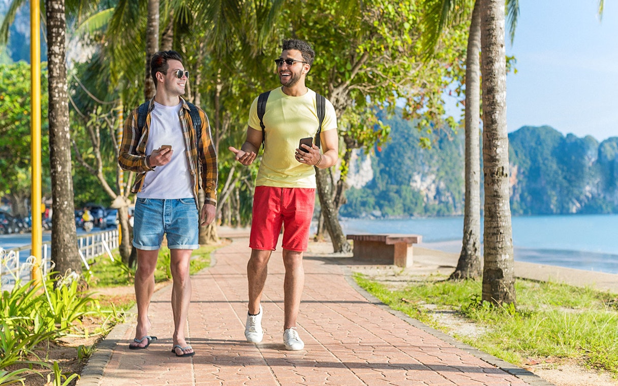 Two people walking on a palm-lined path by the beach, using phones in Kuala Lumpur, Malaysia.