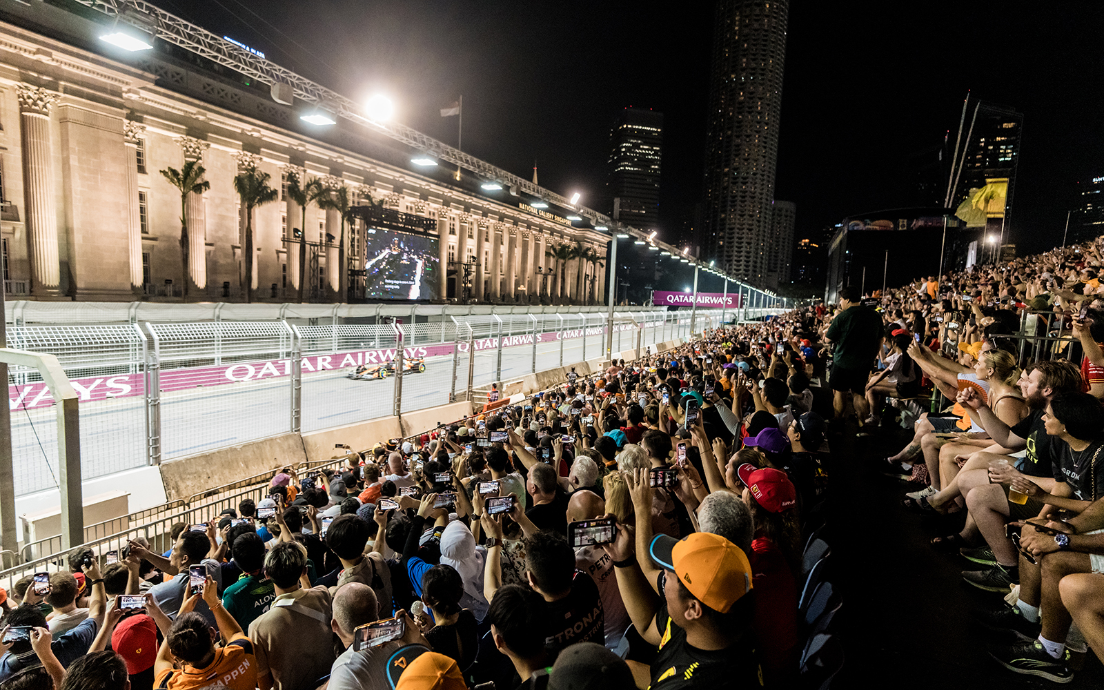 Crowd watching F1 race at night in Singapore with city skyline in background.