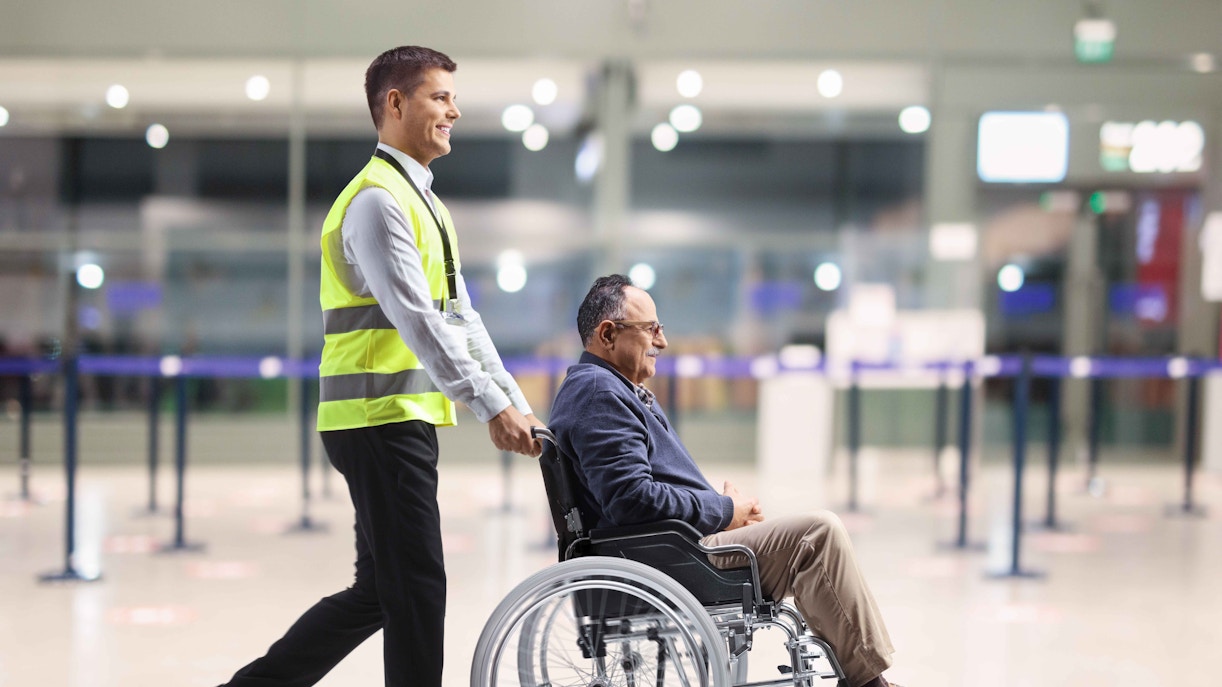 Airport staff assisting passenger in wheelchair through terminal.