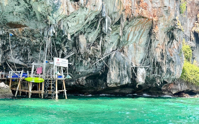 Overwater structures and a kayak docked at the base of a sheer limestone cliff.