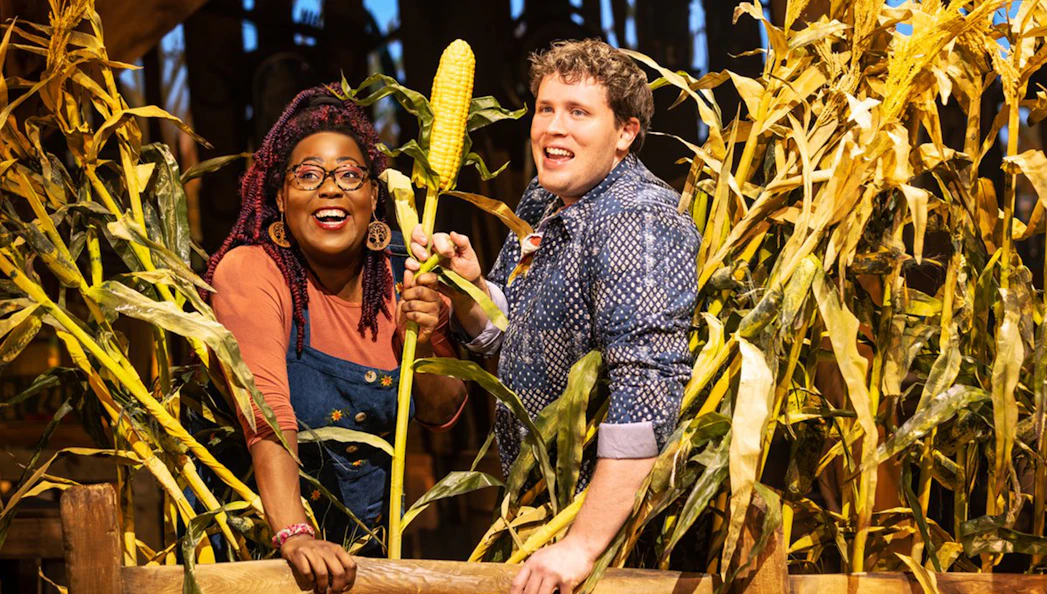 Two people smiling in a cornfield, holding an ear of corn, related to the "Shucked" tour.