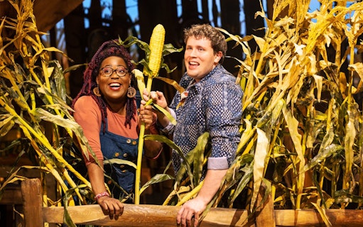 Two people smiling in a cornfield, holding an ear of corn, related to the "Shucked" tour.