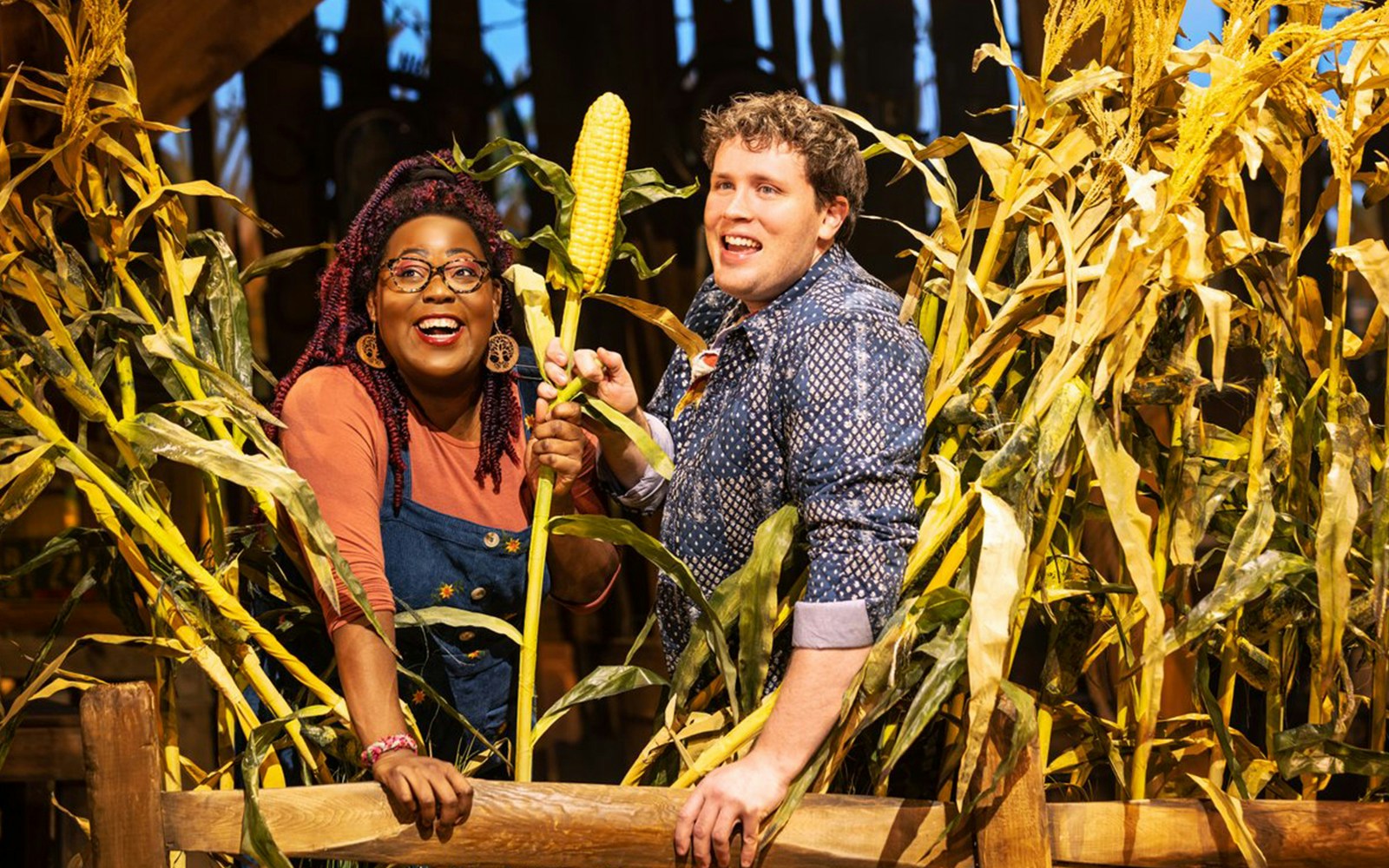 Two people smiling in a cornfield, holding an ear of corn, related to the "Shucked" tour.