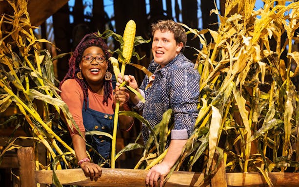 Two people smiling in a cornfield, holding an ear of corn, related to the "Shucked" tour.