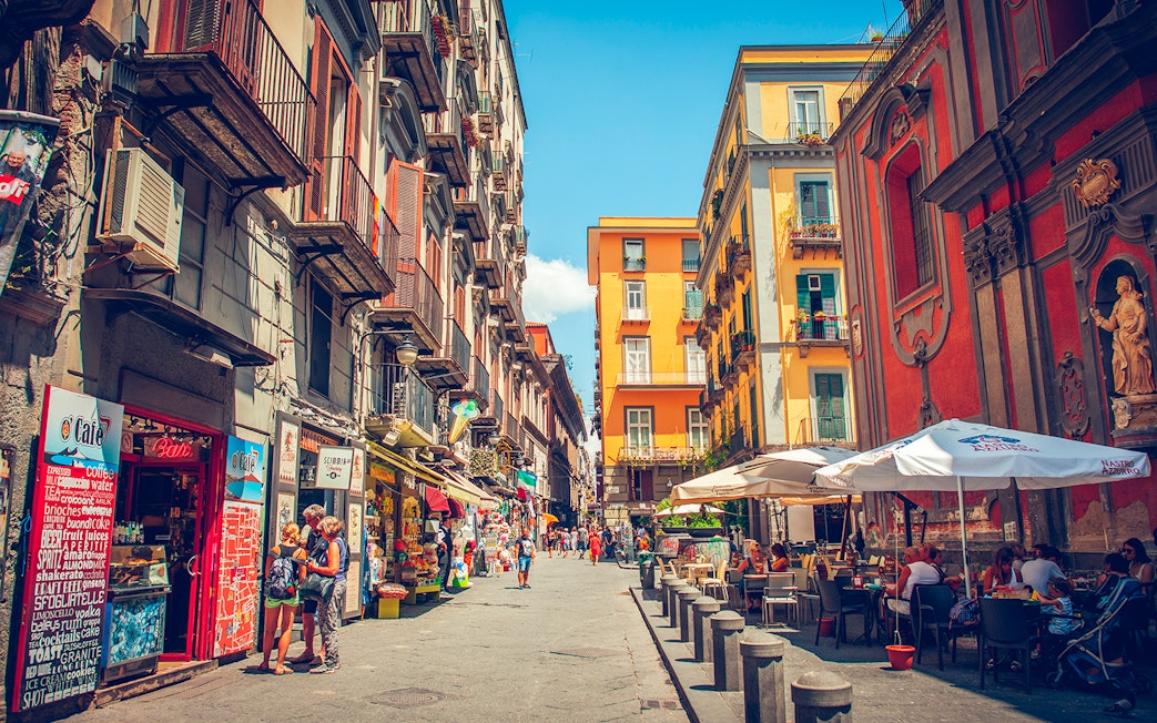 Tourists walking through a vibrant street in Naples with colorful buildings and outdoor cafes.