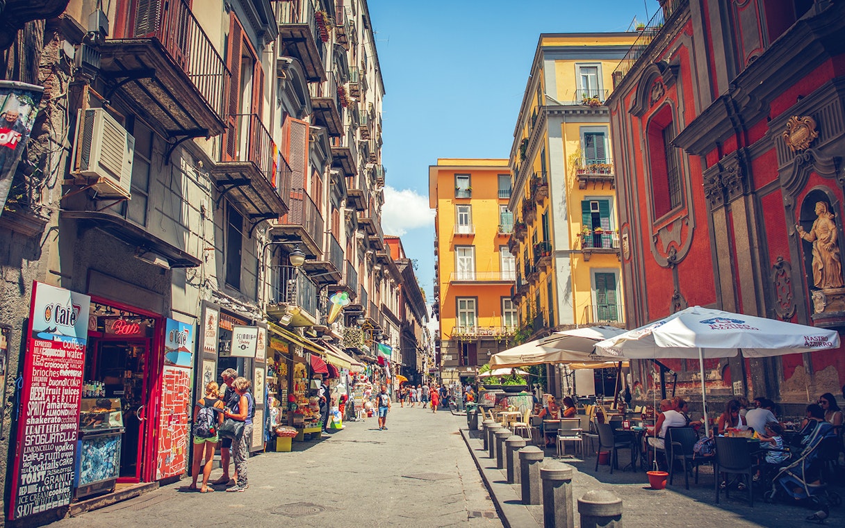 Tourists walking through a vibrant street in Naples with colorful buildings and outdoor cafes.