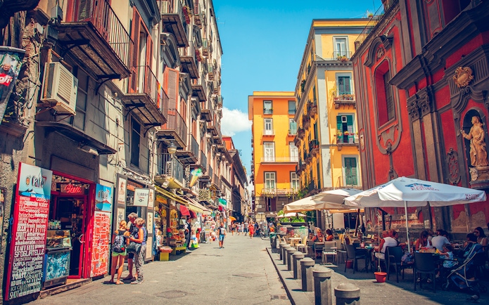 Tourists walking through a vibrant street in Naples with colorful buildings and outdoor cafes.