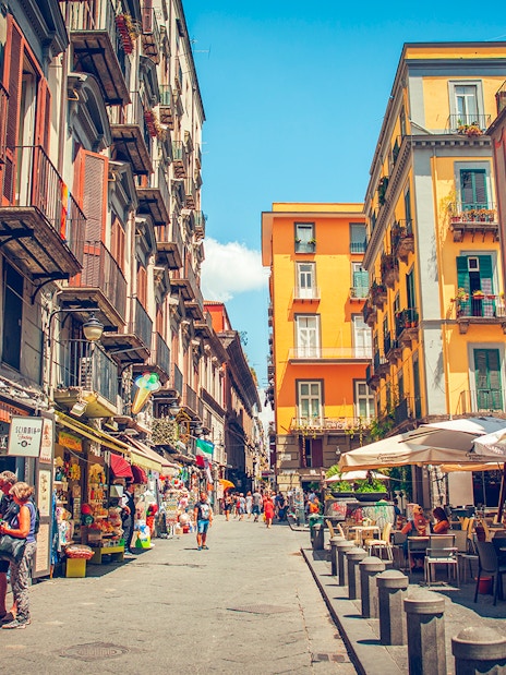 Tourists walking through a vibrant street in Naples with colorful buildings and outdoor cafes.