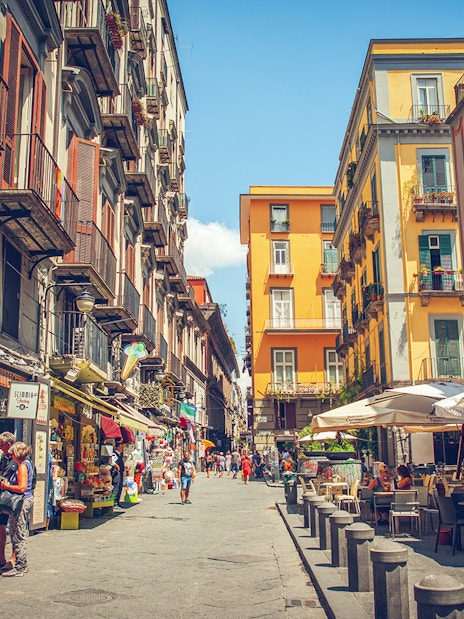 Tourists walking through a vibrant street in Naples with colorful buildings and outdoor cafes.