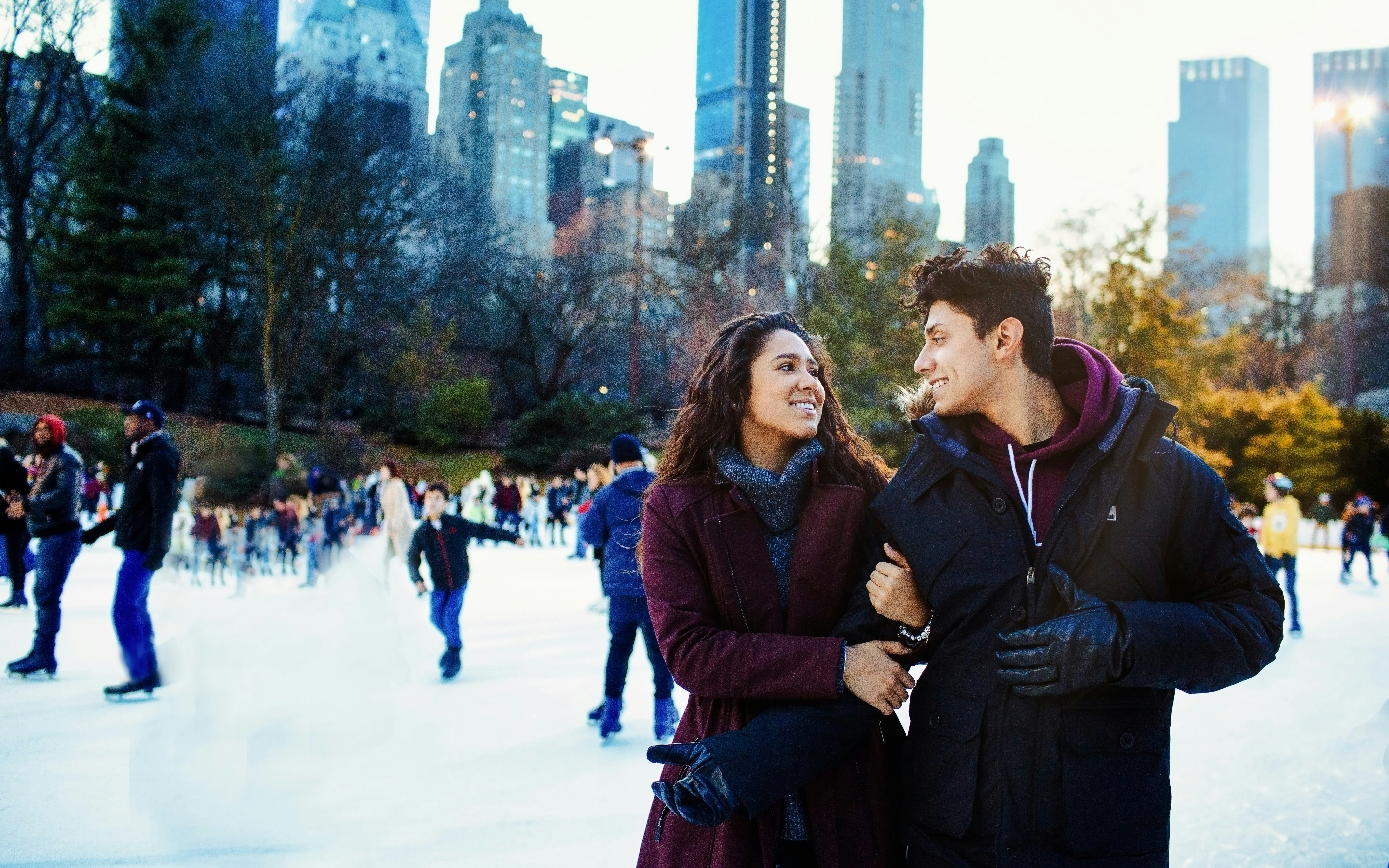 Couple ice skating at Wollman Rink in Central Park, New York City skyline in background.