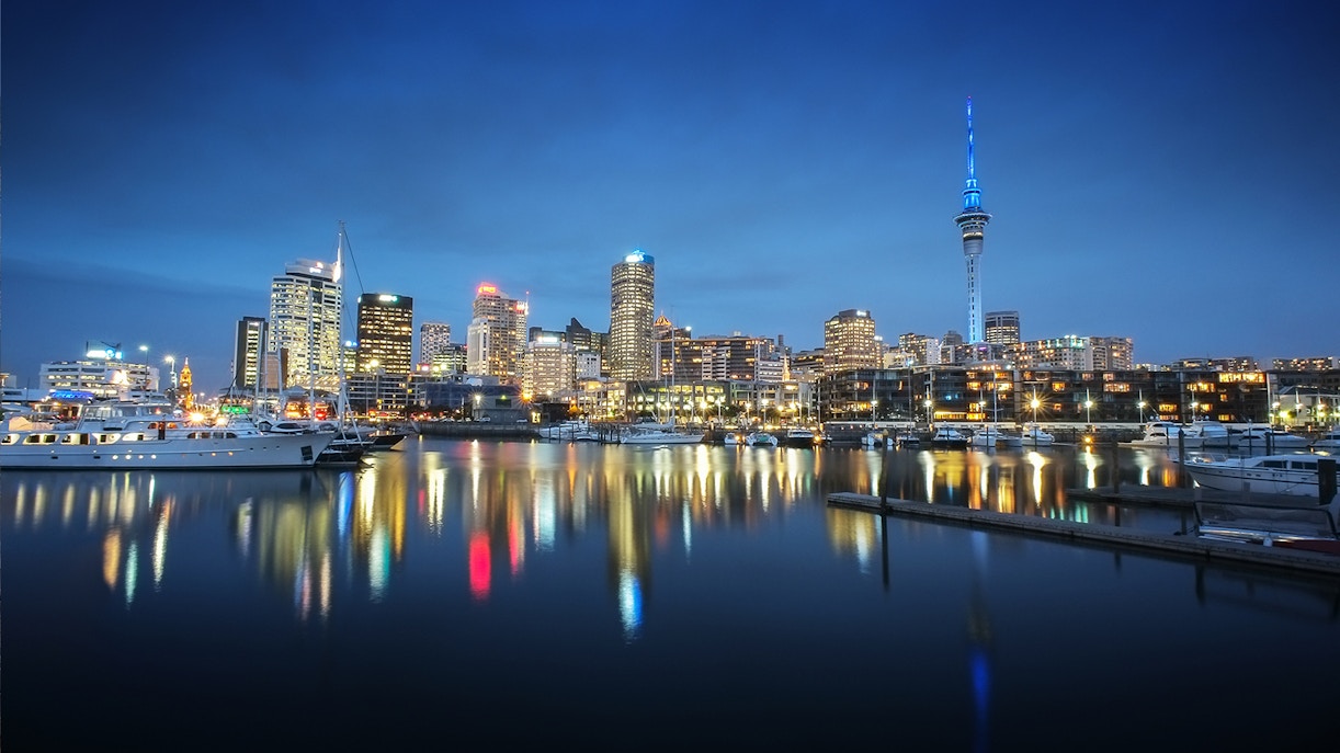 Auckland skyline at night with illuminated Sky Tower reflecting on the harbor.
