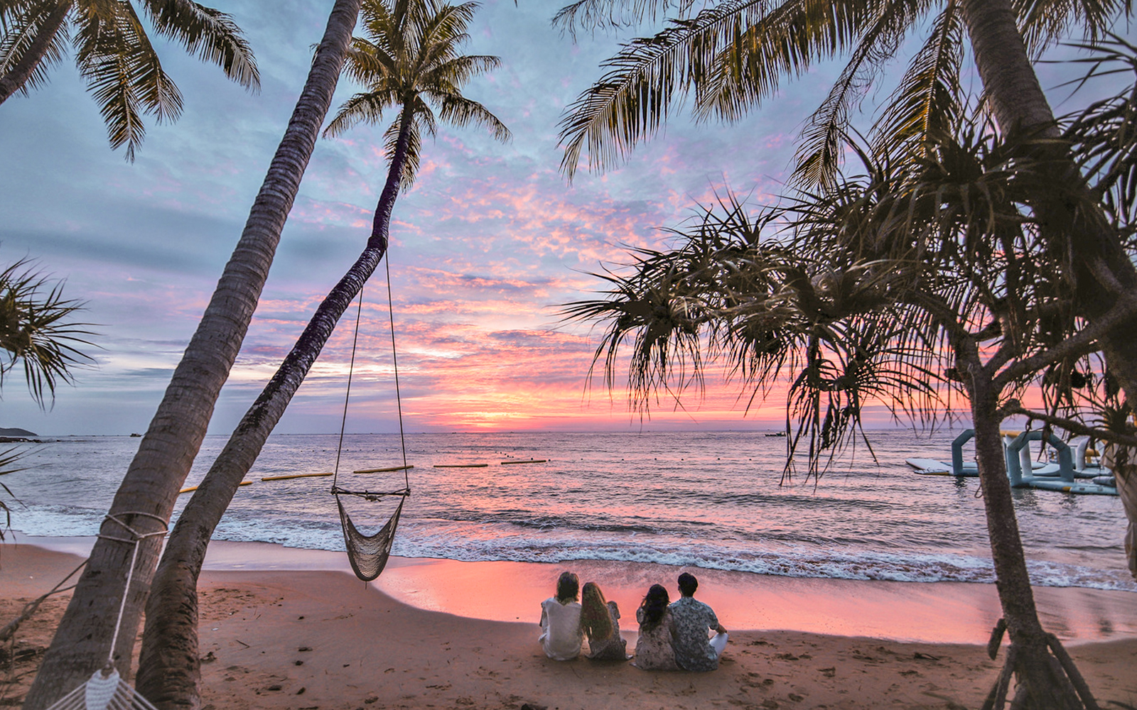Tourists sitting on a beach at sunset with palm trees and ocean view.