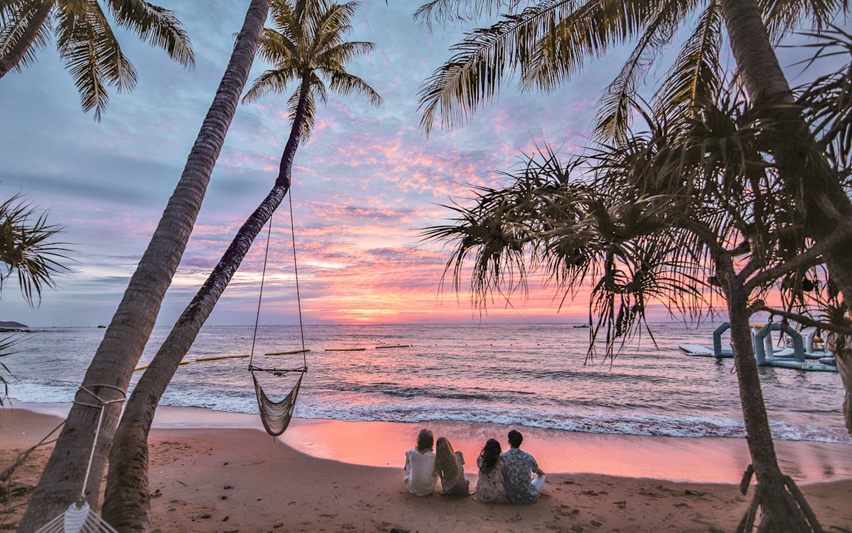 Tourists sitting on a beach at sunset with palm trees and ocean view.