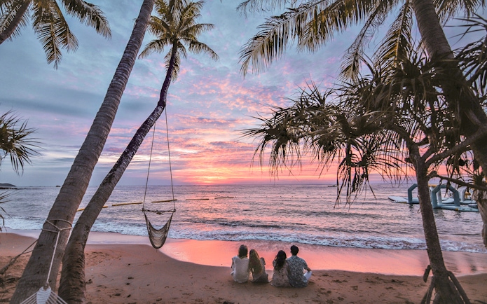 Tourists sitting on a beach at sunset with palm trees and ocean view.