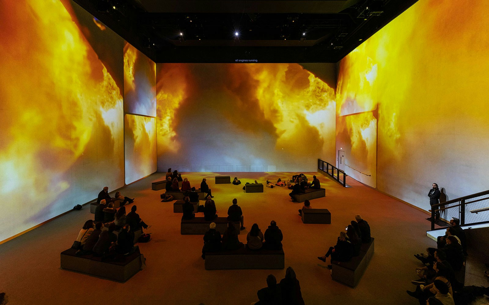 Visitors seated in a large room with immersive fiery visuals at The Moonwalkers experience.
