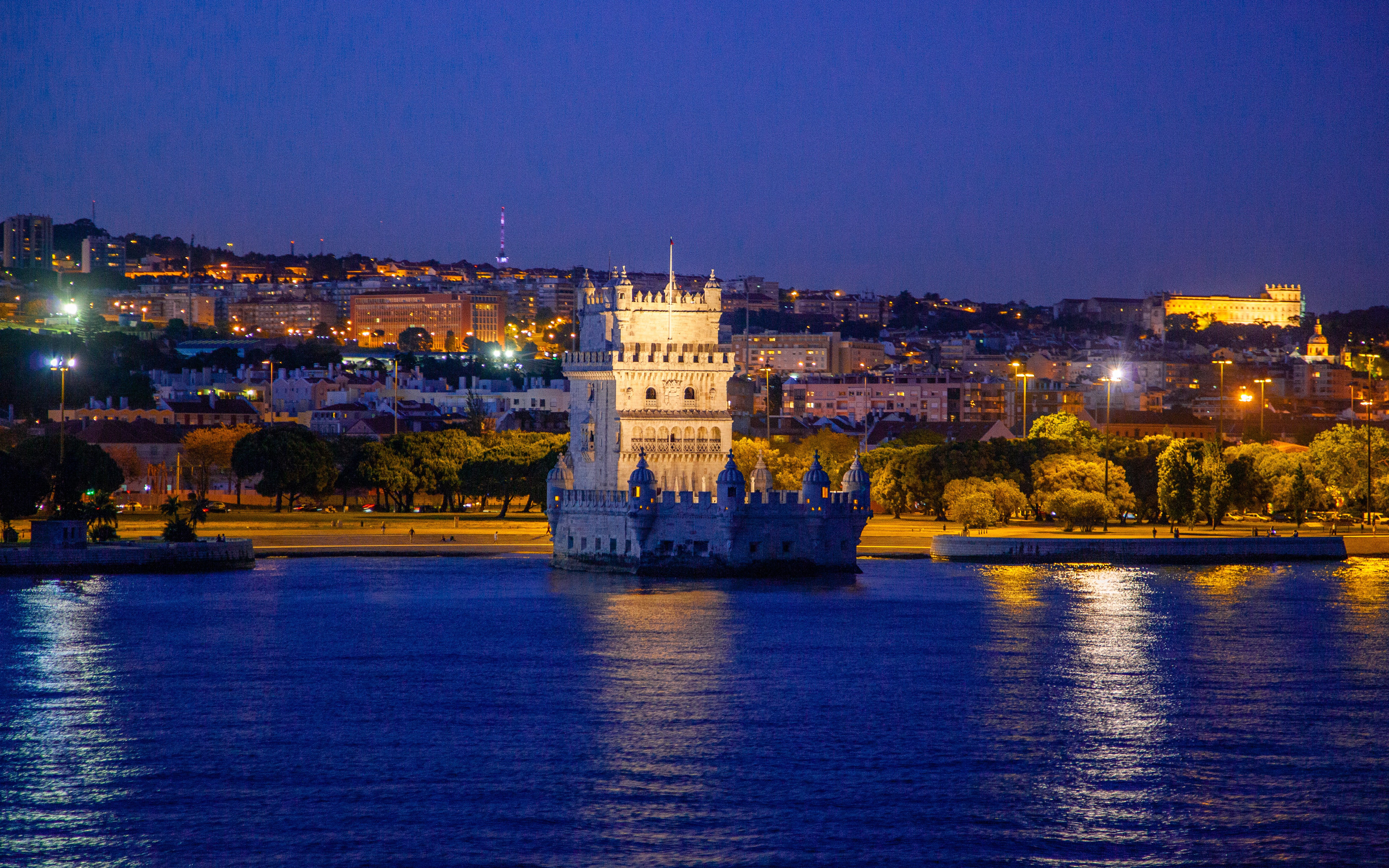 Belém Tower illuminated at night with Lisbon cityscape in the background.