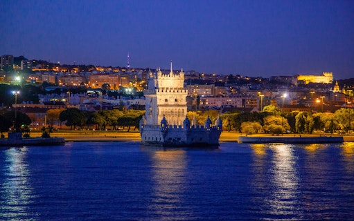 Belém Tower illuminated at night with Lisbon cityscape in the background.