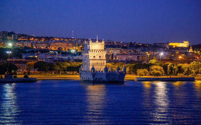Belém Tower illuminated at night with Lisbon cityscape in the background.