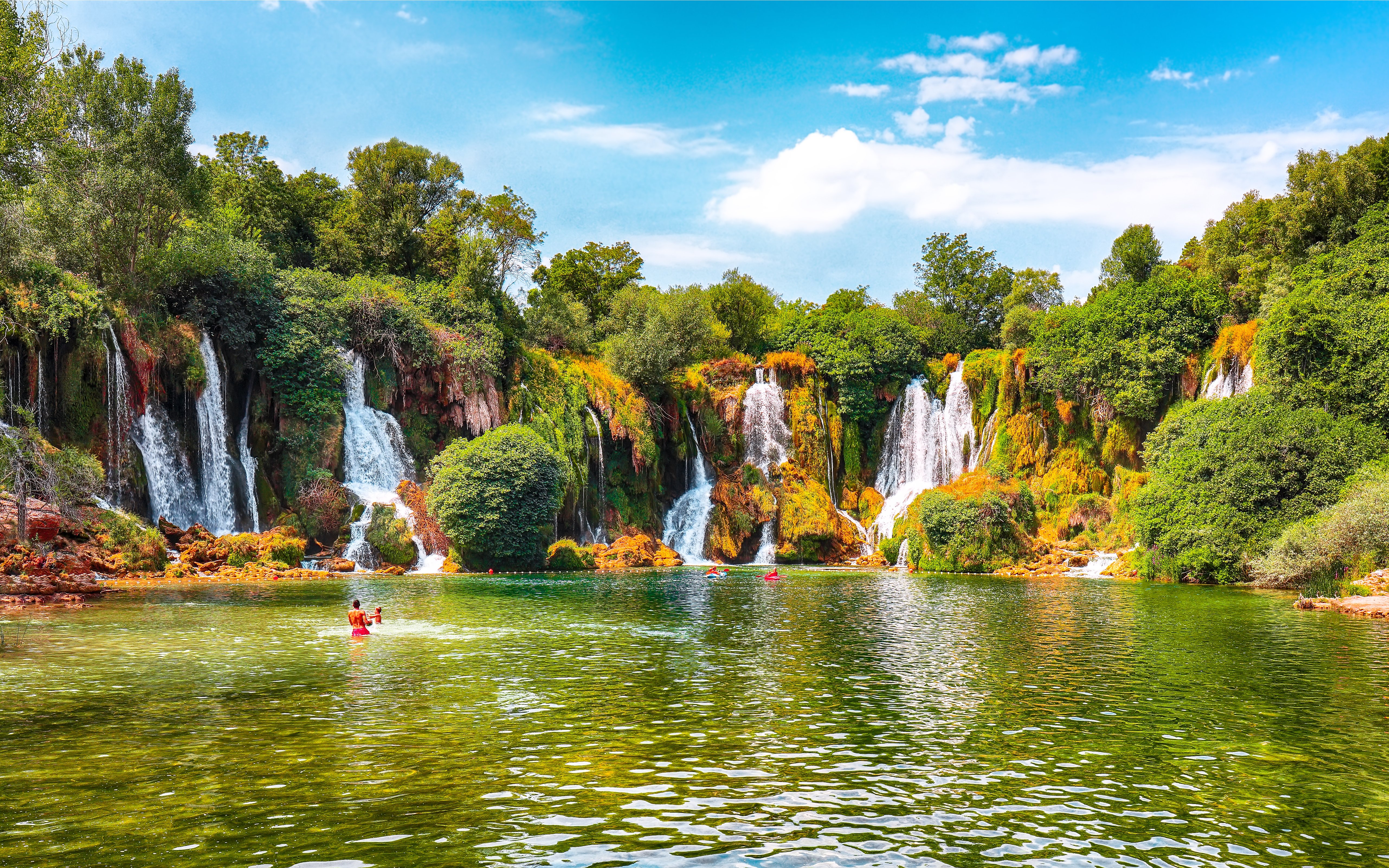 Kravice Waterfall with lush greenery and people swimming in the foreground.