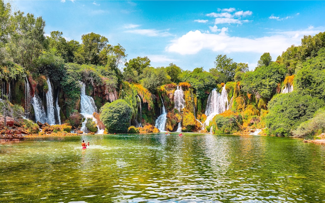 Kravice Waterfall with lush greenery and people swimming in the foreground.