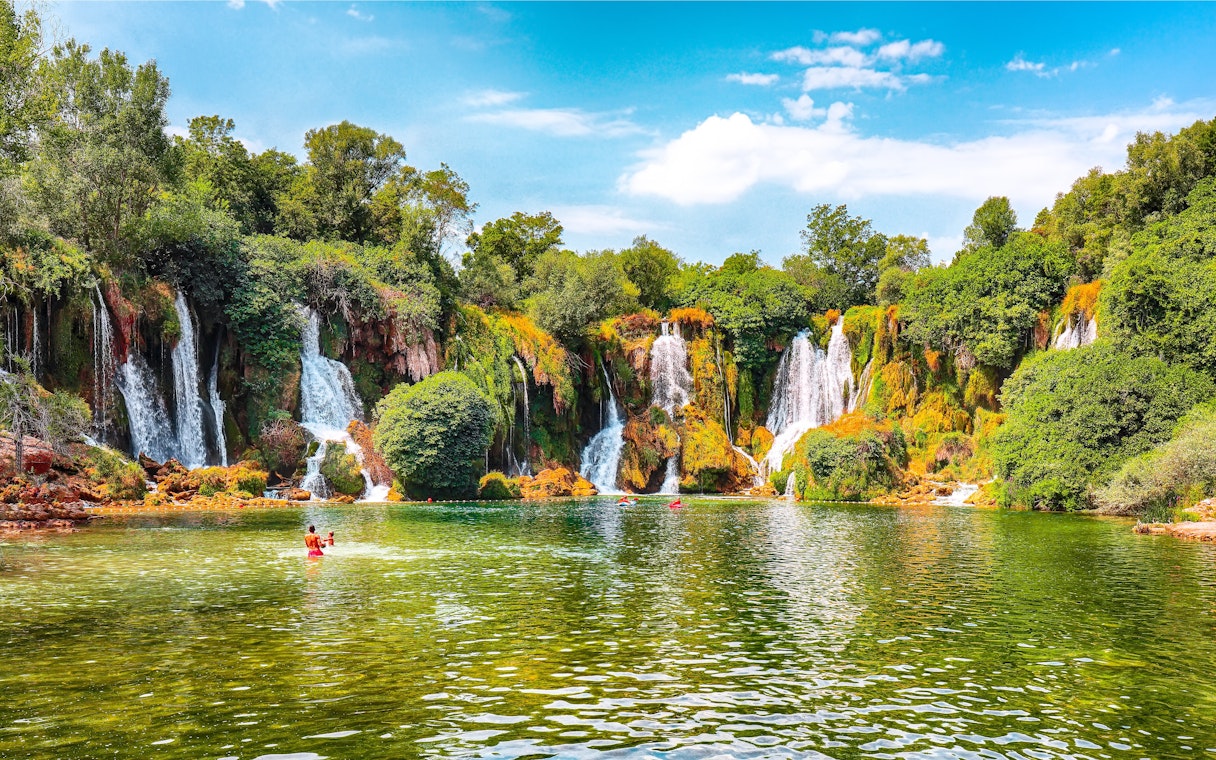 Kravice Waterfall with lush greenery and people swimming in the foreground.